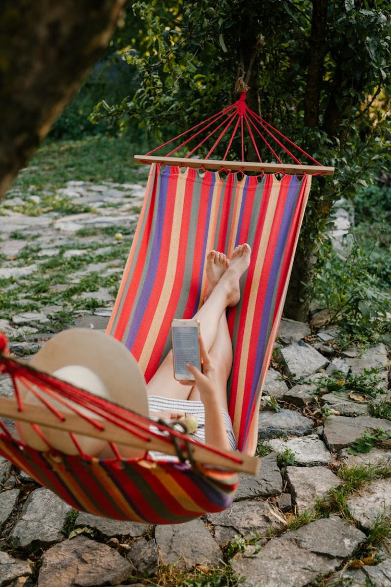 From above full body of anonymous barefoot female lying in hammock and using mobile phone in green garden